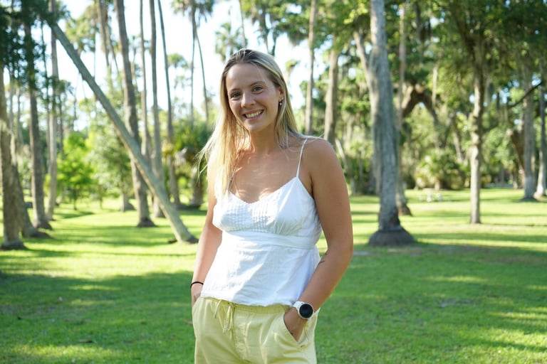 Smiling woman in white tank top and light green pants standing in a sunny park with tall trees