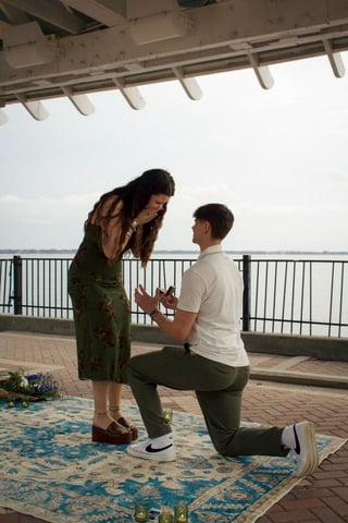 Man on one knee proposing to woman on a waterfront overlook with decorative rug and candles