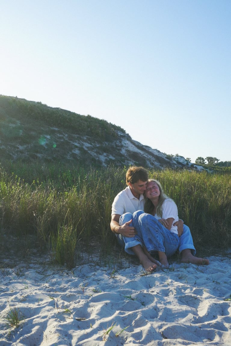 Couple sitting together on sandy beach with dunes and grass in background at sunset