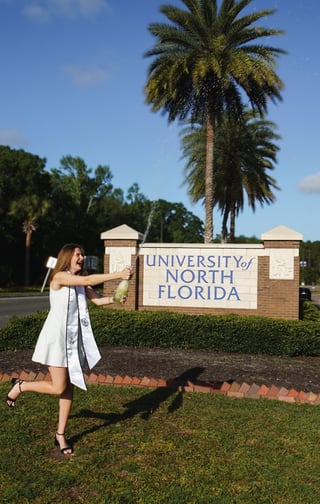 Woman in white dress posing at University of North Florida entrance sign with tall palm tree and brick pillars in background