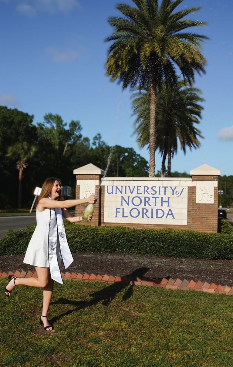 Woman in white dress posing at University of North Florida entrance sign with tall palm tree and brick pillars in background
