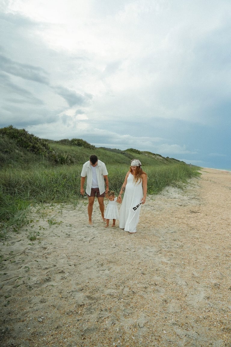 Family of three holding hands walking on sandy beach near dunes under cloudy sky