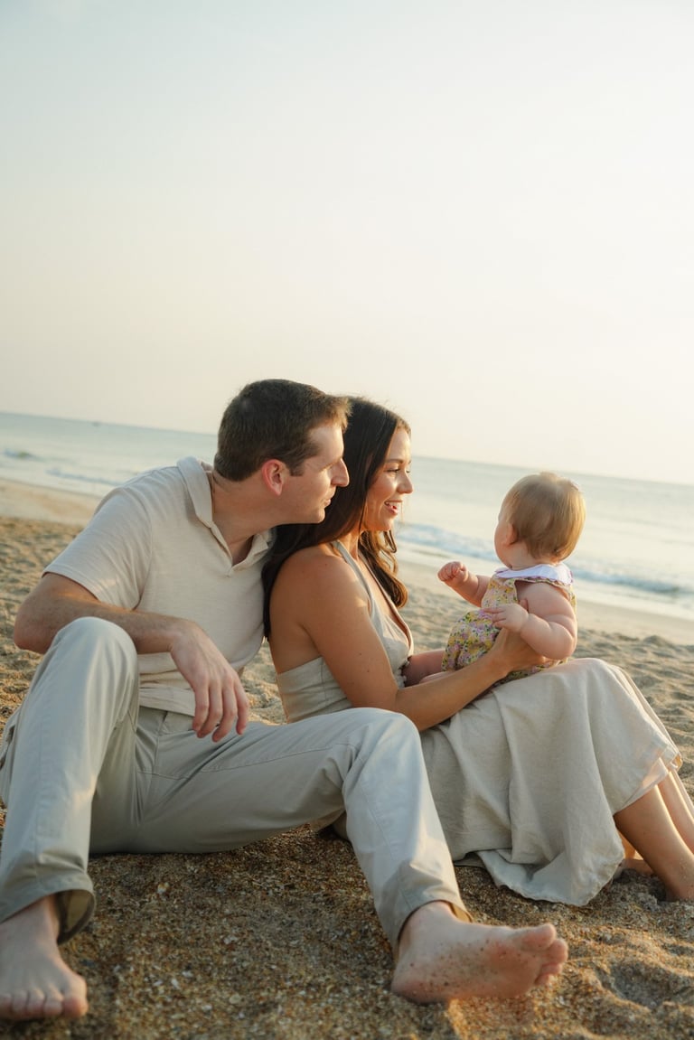 Family of three sitting on sandy beach at sunset, parents in light colored clothing with young toddler
