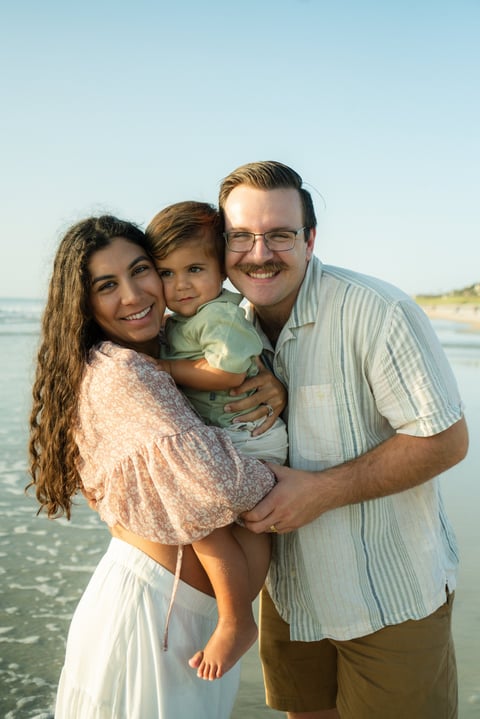 Family of three smiling at the beach, with a father holding a young child between parents, ocean and sky in the background