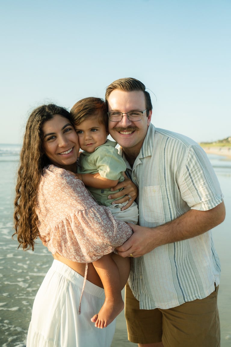 Family of three smiling at the beach, with a father holding a young child between parents, ocean and sky in the background
