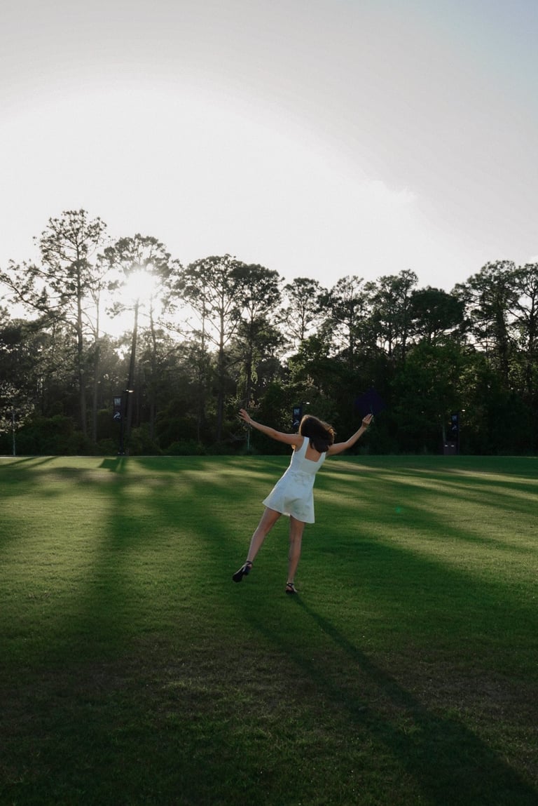 Woman in white dress standing in grassy field with arms raised, surrounded by tall trees under overcast sky