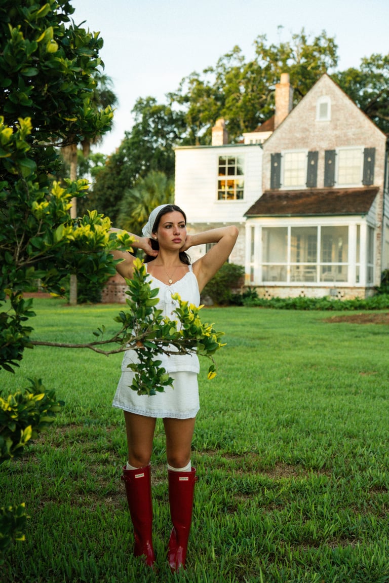 Woman in white dress and red boots standing in front of farmhouse with green lawn and flowering trees