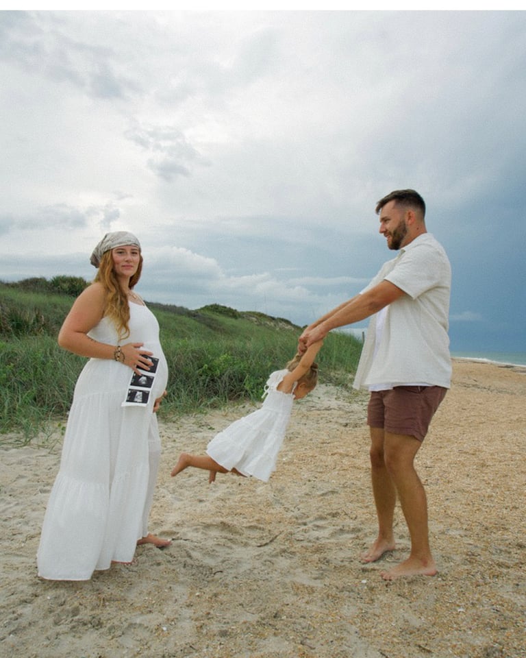 Pregnant couple playing with toddler on beach, holding child up by hands against cloudy sky and dunes